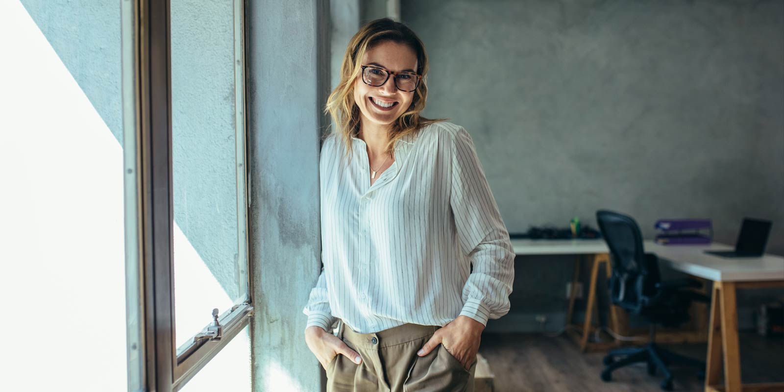 Frau mit Brille und gestreifter Bluse lehnt lächelnd an einer Wand in einem modernen Büro.