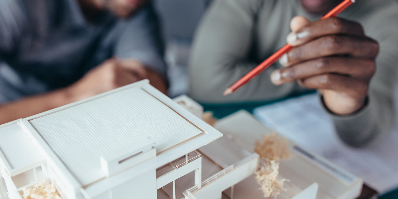 Zwei Personen begutachten ein Architekturmodell; eine Hand mit rotem Bleistift zeigt auf das Modell.